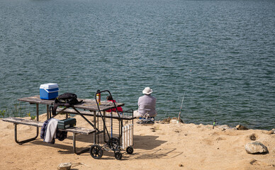 Winchester, CA, USA - April 11, 2022: Skinner Lake. In front of dark blue-gray water, Fisher sits, back to us, on sandy rocky shoreline with his tools exposed on old wooden bench.