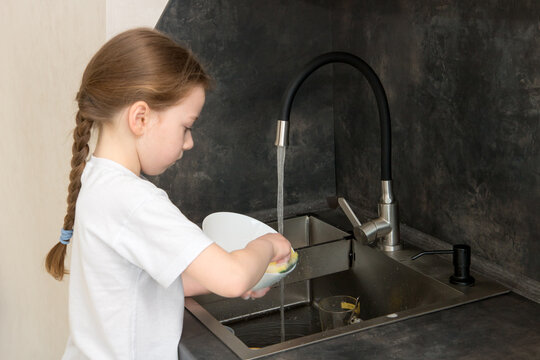 cute little girl with a pigtail washes the dishes in the kitchen at the sink