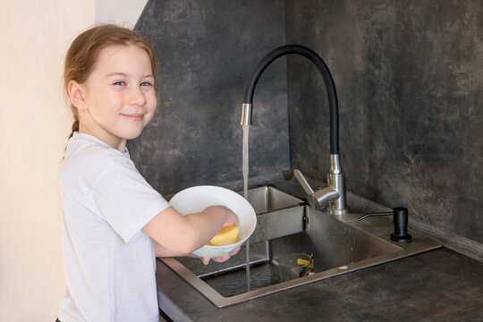 cute little girl with a pigtail washes dishes in the kitchen at the sink and smiles