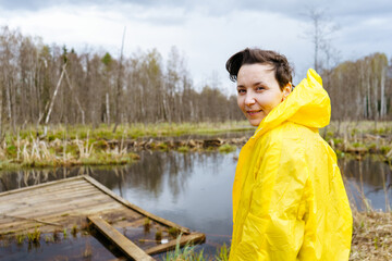 A young woman in a yellow raincoat stands by a lake in the forest and looks at the camera