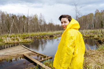 A young woman in a yellow raincoat stands by a lake in the forest and looks at the camera