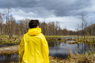 A young woman in a yellow raincoat stands next to a lake and looks at the landscape with her back to the camera