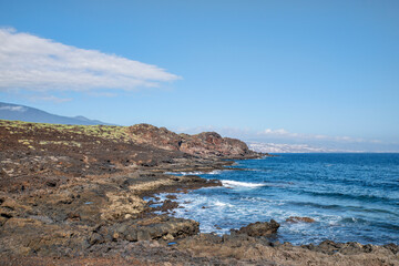 Path through the rough volcanic lands known as malpais, at Puertito de Guimar, with clear ocean waves and views of mountain range and El Socorro town in the distance, Tenerife, Canary Islands, Spain