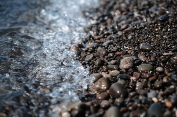 Small stones in azure sea water close-up. A picture of the sea surf. Ocean surf among the rocks. Background of the tide. Sea foam