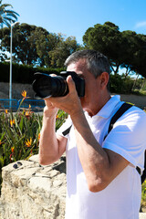 A 60 year old man wearing sunglasses, on summer vacation sightseeing in the city of Alicante, taking pictures with a camera in front of the Santa Barbara Castle.