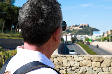  A 60 year old man wearing sunglasses, on summer vacation sightseeing in the city of Alicante, looking at the Santa Barbara Castle.