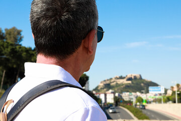  A 60 year old man wearing sunglasses, on summer vacation sightseeing in the city of Alicante, looking at the Santa Barbara Castle.