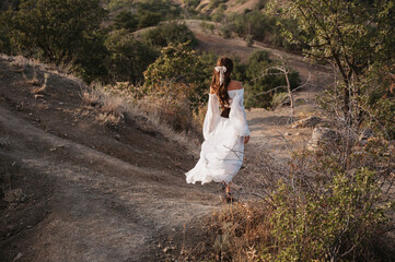 A young girl in a long white dress is walking down a path in a hilly area among trees. The concept of freedom and travel.