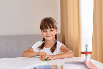 Naklejka premium Portrait of of dark haired little girl drawing something in album sitting at the table near window, cute kid with braids looking at camera with happy facial expression.
