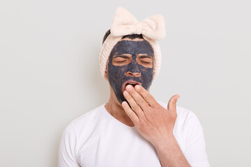 Portrait of funny man wearing white t shirt and female's hair band standing with mud mask and yawning, covering mouth with palm, being tired to wait result posing isolated over gray background.