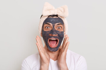 Indoor shot of funny excited surprised man wearing white t shirt and hair band with bow posing isolated over gray background, screaming with mud mask on face and raised arms.