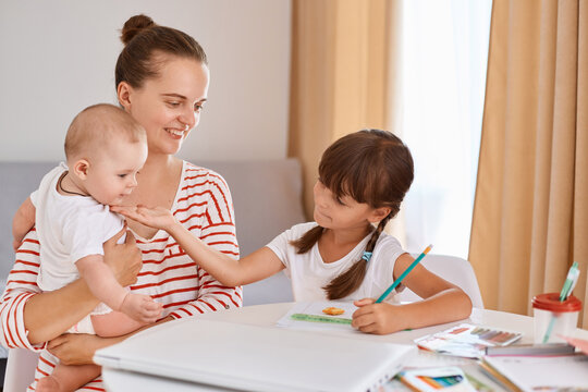 Indoor shot of smiling woman holding infant baby and helping her elder daughter with homework, sitting at table and writing, posing in light living room.