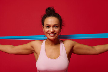 Fototapeta na wymiar Close-up of a charming determined sportswoman exercising with elastic fitness band, stretching her arms against red background with copy ad space