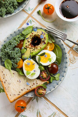 Healthy lunch with avocado halves, cherry tomatoes, soft-boiled eggs, kale, wheat bread, seeds and soy sauce flavored with edible flowers.