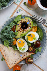 Healthy lunch with avocado halves, cherry tomatoes, soft-boiled eggs, kale, wheat bread, seeds and soy sauce flavored with edible flowers.