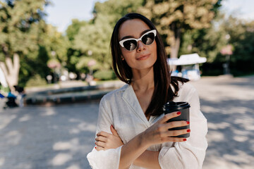 Elegant stylish woman with straight dark hair wearing sunglasses and white shirt is holding cup with coffee and posing at camera in sunlight in green summer park in the city