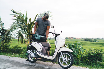 Fototapeta na wymiar Young male tourist with backpack standing near vintage scooter parked near tropical palm and path way, casual dressed African American hipster guy traveling on retro moped during trip in Indonesia