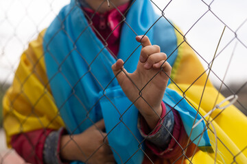 sad little girl with the flag of Ukraine behind a metal fence. Social problem of refugees and...