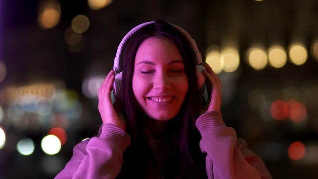 A beautiful brunette girl in white headphones listens to music and smiles under a purple light on a city street at night. Portrait
