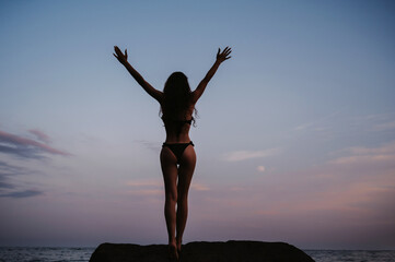 Silhouette of a beautiful young girl with a sexy booty standing on the rocks on the beach by the...