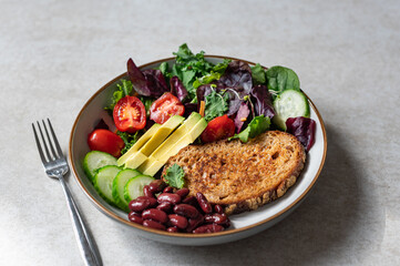 Light green salad with leaves, avocado, cherry tomatoes, cucumber slices, red beans and slice of toasted bread