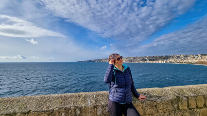 A tourist woman enjoying the panoramic view from Castel dell Ovo (Egg Castle) on the city of Naples, Campania, Italy, Europe. Person leaning against the castle wall, bathing in the sun. Sea view