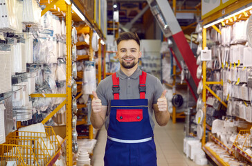 Happy salesman at modern DIY store guarantees best quality of all the goods. Portrait of handsome young man in uniform standing between shelves with ventilation grids, smiling and showing thumbs up