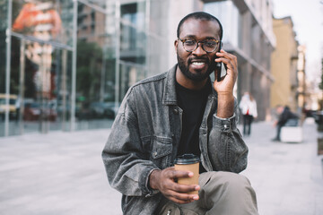 Portrait of dark skinned man in classic eyewear looking at camera while cell talking in urban city, millennial African American hipster guy with takeaway coffee using mobile technology for calling