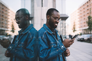 Happy dark skinned blogger in classic spectacles browsing web content and smiling during time in city, cheerful hipster guy using mobile application for checking internet connection on cellphone