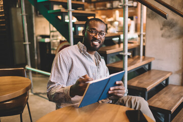 Portrait of happy student in eyewear holding copybook for creating education essay story, cheerful African American male with notebook for organization - learning and studying in coworking space
