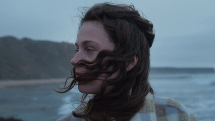 Closeup shot of pretty teenage girl with long dark curly hair wearing casual cosy plaid shirt, staying alone, thinking, dreaming, watching at camera, catching wind and breeze near grey ocean seashore