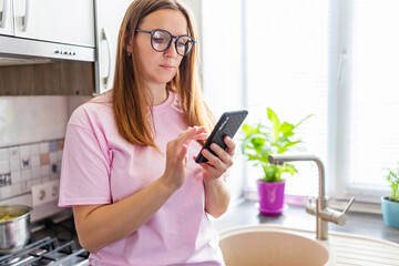 Fototapeta na wymiar Beautiful young woman in eyeglasses using mobile phone at home kitchen. Brunette girl reading or typing a text message