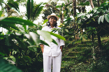 Portrait of successful Balinese male in hat looking at camera during daytime at seedling field with coffee bush, adult farmer posing while visiting countryside caffeine plantation in Indonesia