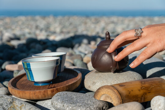 Traditional Chinese tea ceremony on the pebble beach of the sea in sunny weather. Close-up of a woman touching a clay teapot with her hand, there are two cups on a wooden tray nearby