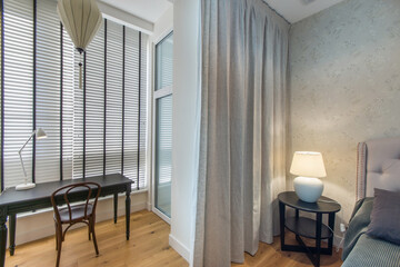 A modern bedroom in bright colors with a table by the window with blinds. The bedside table is decorated with a lamp.