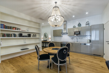 A spacious room in bright colors with a luxurious chandelier, a shelf, a kitchen and a dining table with chairs. The floor is covered with a brown parquet board.