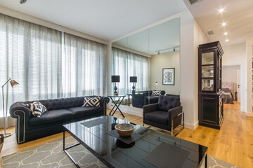 Modern interior of the living room with a large window and a mirrored wall. A dark glass coffee table and a gray sofa with pillows.