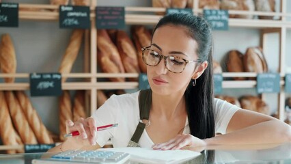 Video of beautiful woman seller checking stock of pastry while counting the benefits of the day with the calculator in the pastry shop.