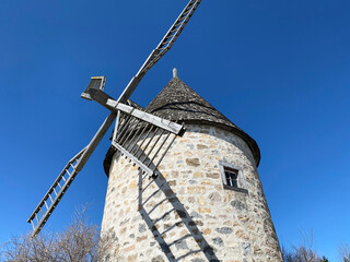 Moulin à vent en pierre des champs avec une toiture en bois. Palmes du moulin à moudre sous sur un ciel bleu. Tour en briques avec une petite fenêtre en bois. © Stephanie