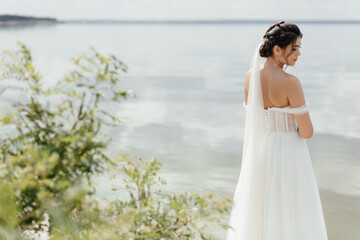 the bride in a white wedding dress stands with her back to the lake among sedges and trees