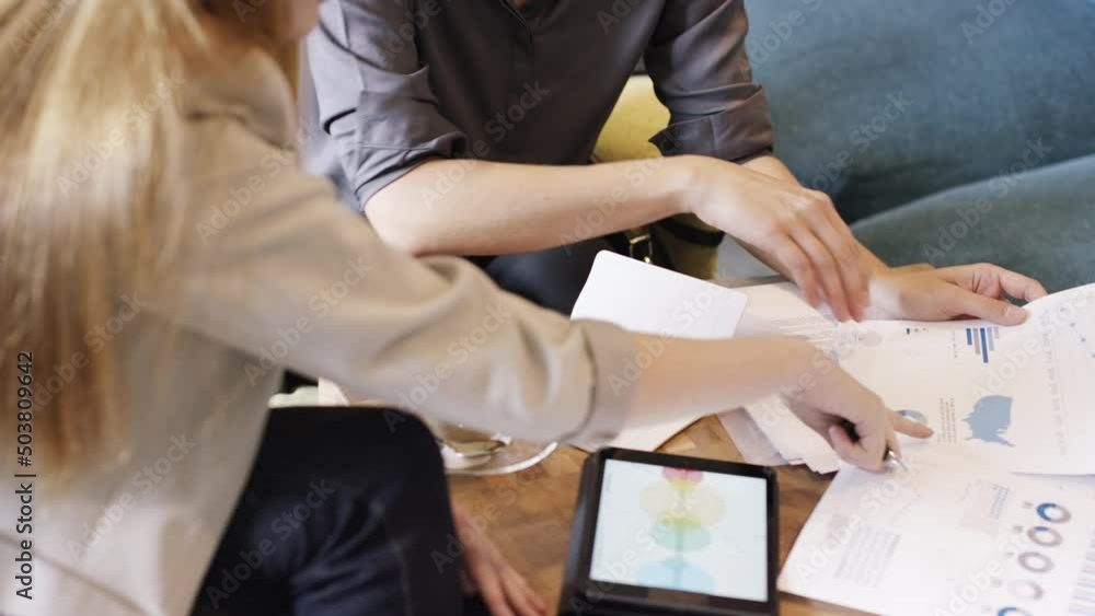 Poster Two businesswomen reading finance documents during a meeting while using a wireless tablet. Two business colleagues collaborate and plan during a meeting while talking and reading documents in a cafe