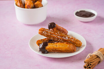 Churros, Spanish choux pastry sweet, deep fried, on a white plate on a pink concrete background. Served with chocolate sauce. Sweets recipes.