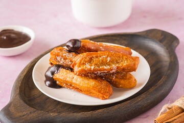 Churros, Spanish choux pastry sweet, deep fried, on a white plate on a pink concrete background. Served with chocolate sauce. Sweets recipes.
