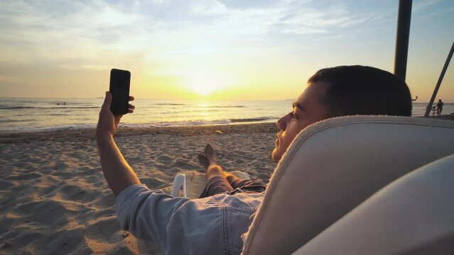 Young enjoying on the lounge chair on the sany tropical beach at sunset, sunrise and taking a selfie with smart phone, vacation and summer travel