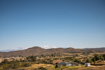 Temecula, CA, USA - April 23, 2022: Snow covered San Jacinto mountains viewed from rural area north of Temecula under blue sky, with brown hills and yellow farmland up front.