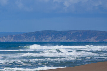 Fototapeta premium Crashing waves with mist rolling in to shore beautiful image for background with hills and bright blue sky