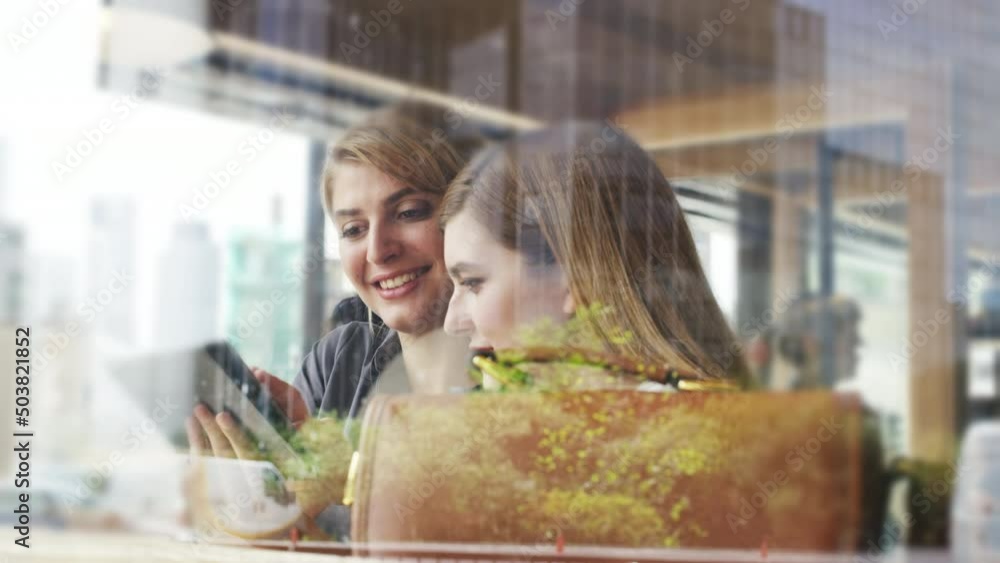 Poster two cheerful businesswomen sitting by a window in a cafe talking and laughing while using a wireless