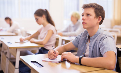 Teen boy sitting at desk in classroom full of pupils during lesson