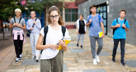 Cute teen girl in glasses walking outside college building in warm autumn day, going to lesson.