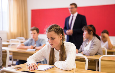 Portrait of positive teenage female pupil sitting at desk studying in classroom